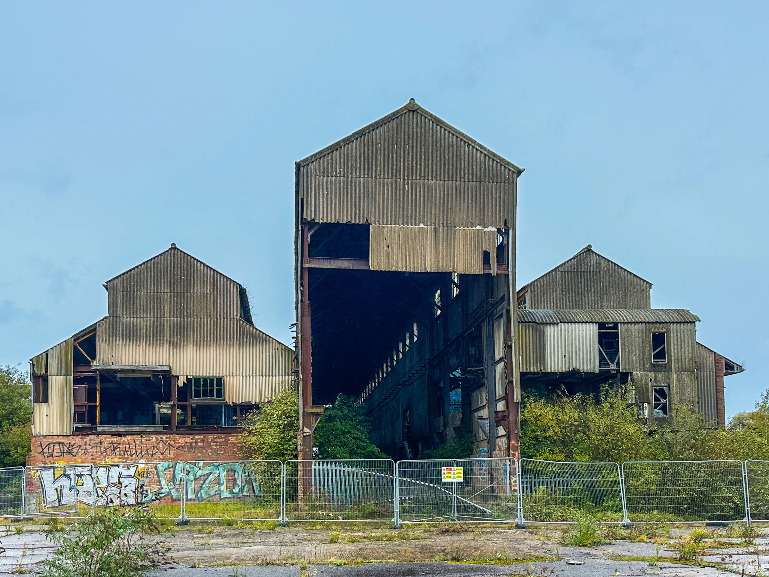 Three gabled corrugated-iron buildings, gutted and open to the weather, fenced off behind metal barriers