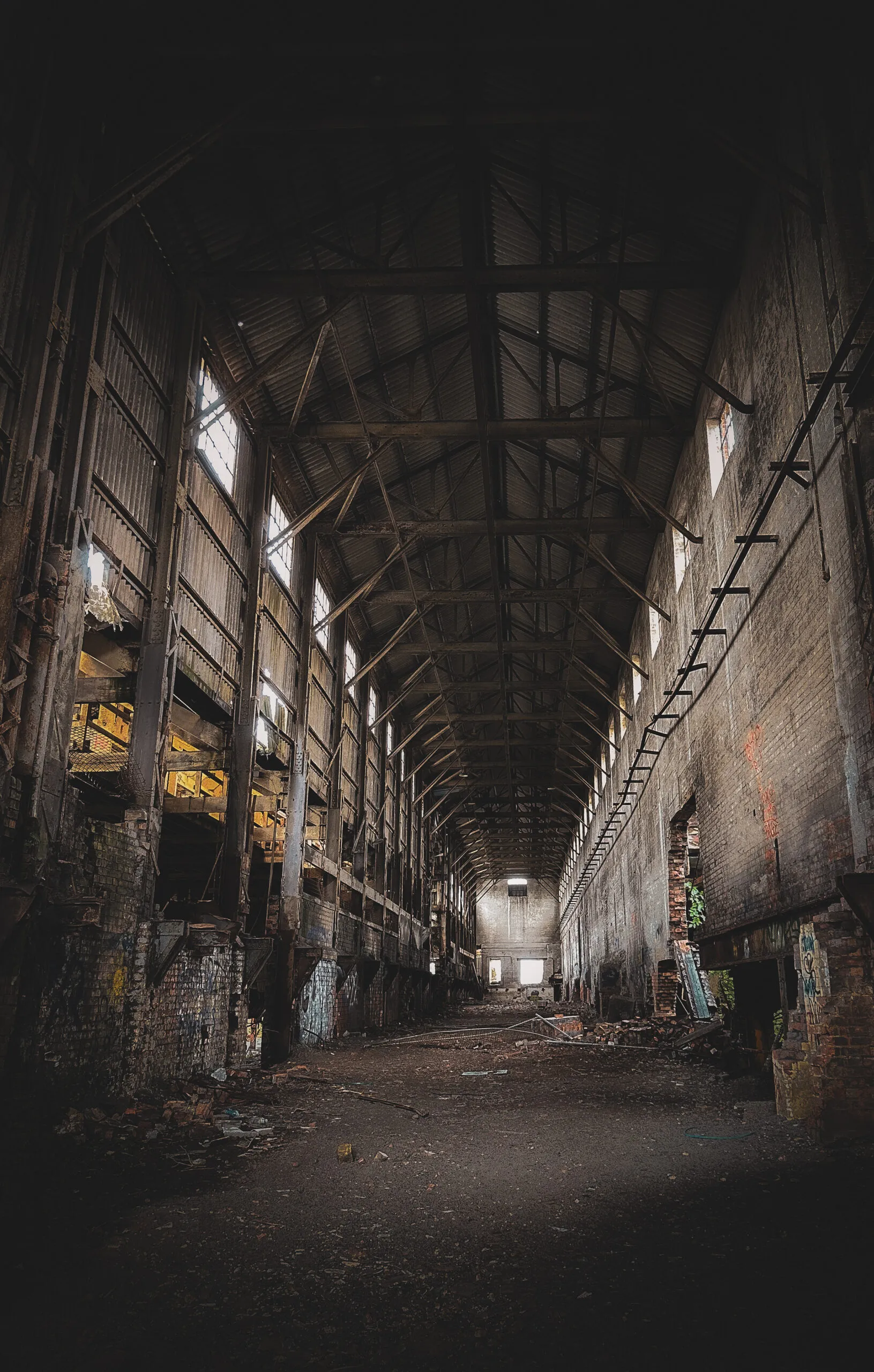 A long, dark brickworks hall receding towards a distant lit window, roof trusses overhead and rubble along the floor