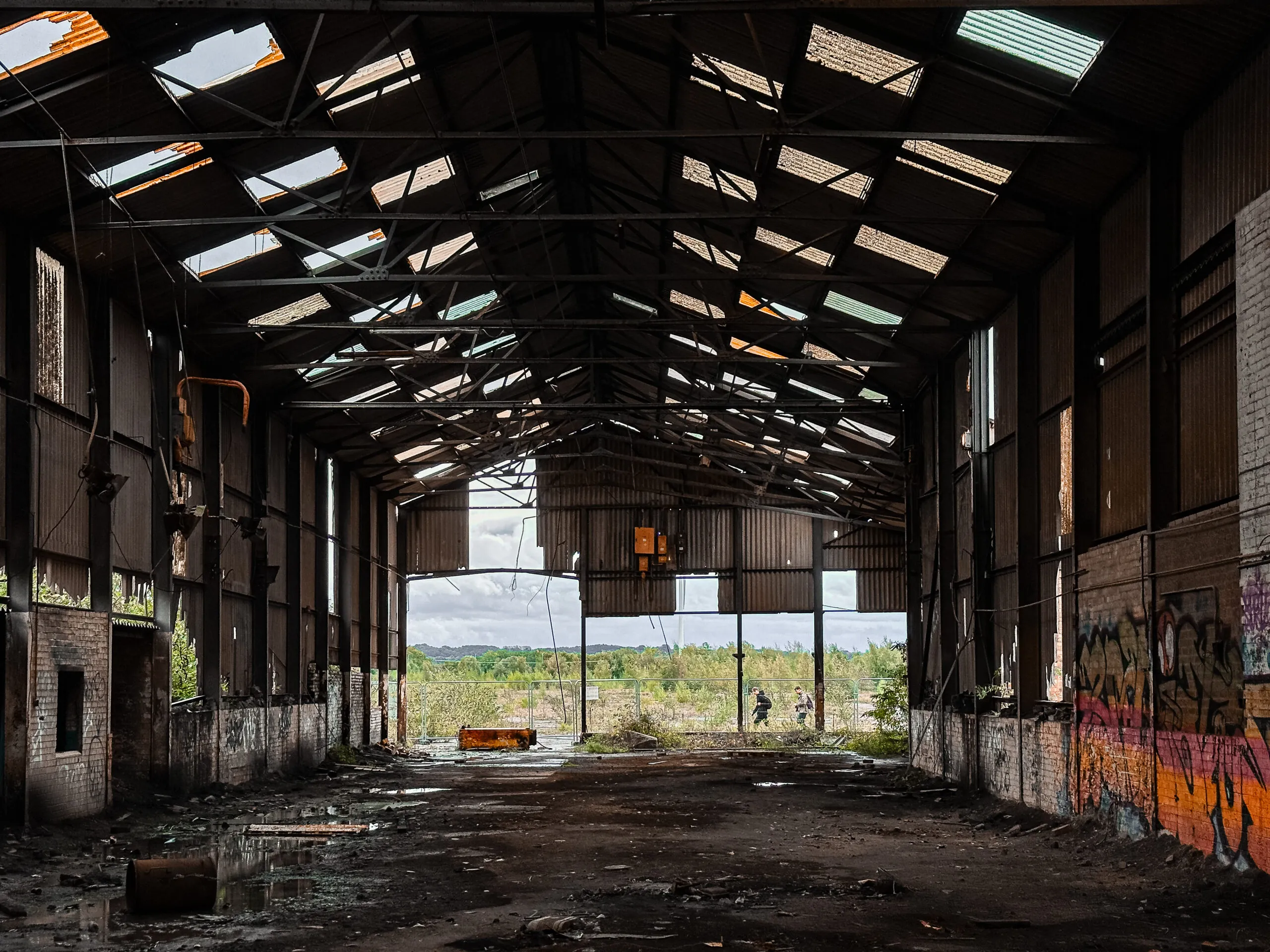 Inside a derelict production hall, daylight falling through a shattered skylight roof onto a wet floor, graffiti along one wall