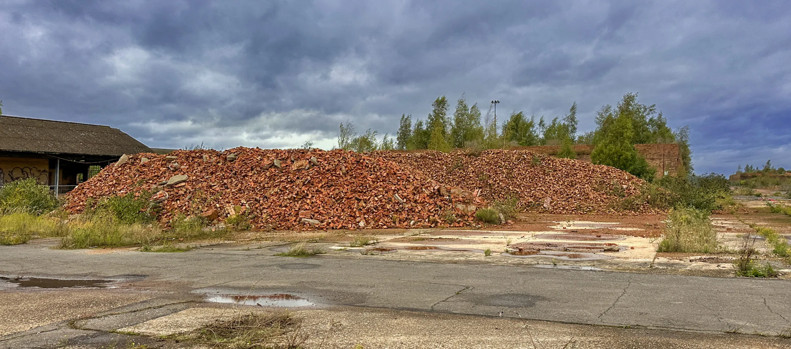A long mound of broken red brick rubble heaped across cracked concrete hardstanding under a heavy storm sky