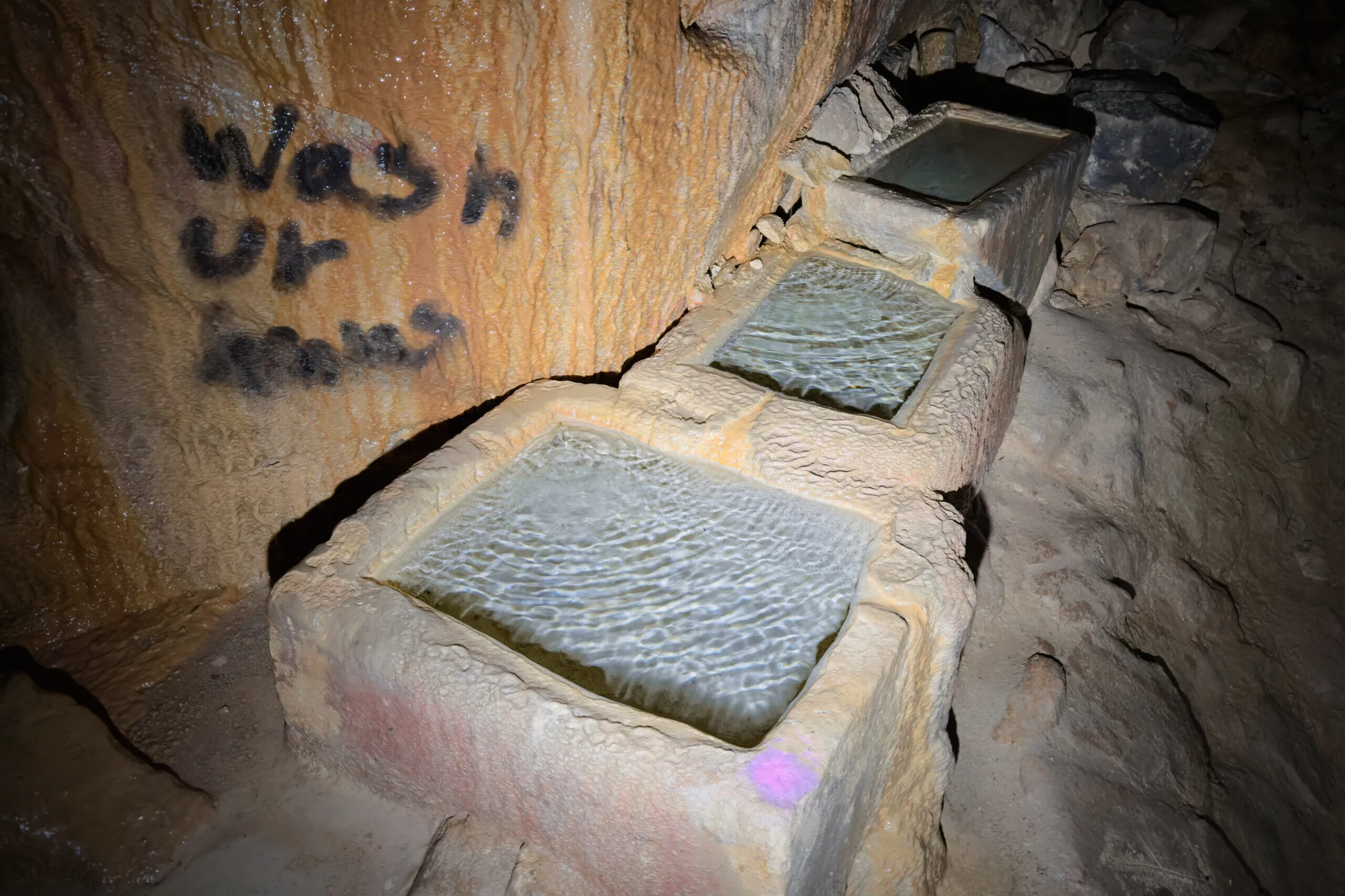 Three stone troughs brimming with clear water, 'wash ur hands' sprayed on the rock above them